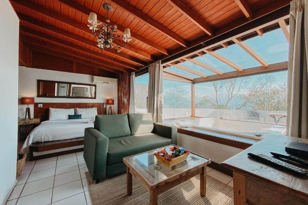 Quarto rústico da Pousada Le Siramat com uma cama de casal, uma janela com vista para as montanhas, todos os móveis são de madeira, o espelho, um sofá, as mesinhas de cabeceira e uma mesa de centro.