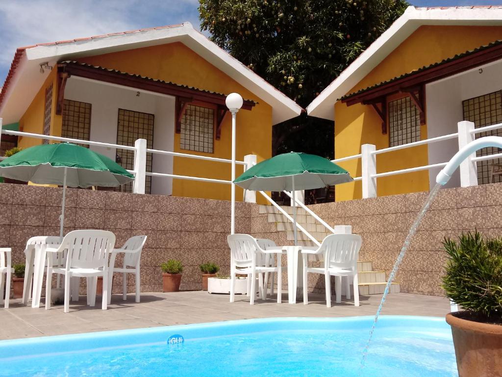 Uma piscina com chafariz no Villa Manary. No deque podemos ver mesas e cadeiras de plástico com guarda-sóis. Atrás, há uma escada para subir e acessar os quartos da pousada.