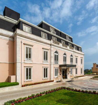 Propriedade do Olissippo Lapa Palace – The Leading Hotels of the World com um prédio rosa clarinho com telhado preto, o local é de época com três andares, na frente há um jardim, para representar hotéis de charme em Lisboa