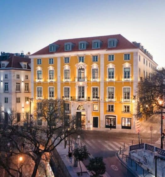 Propriedade do Palácio Ludovice Wine Experience Hotel, uma construção amarela de época com estilo português, com quatro andares, janelas brancas, em uma esquina iluminada, para representar hotéis boutique em Lisboa