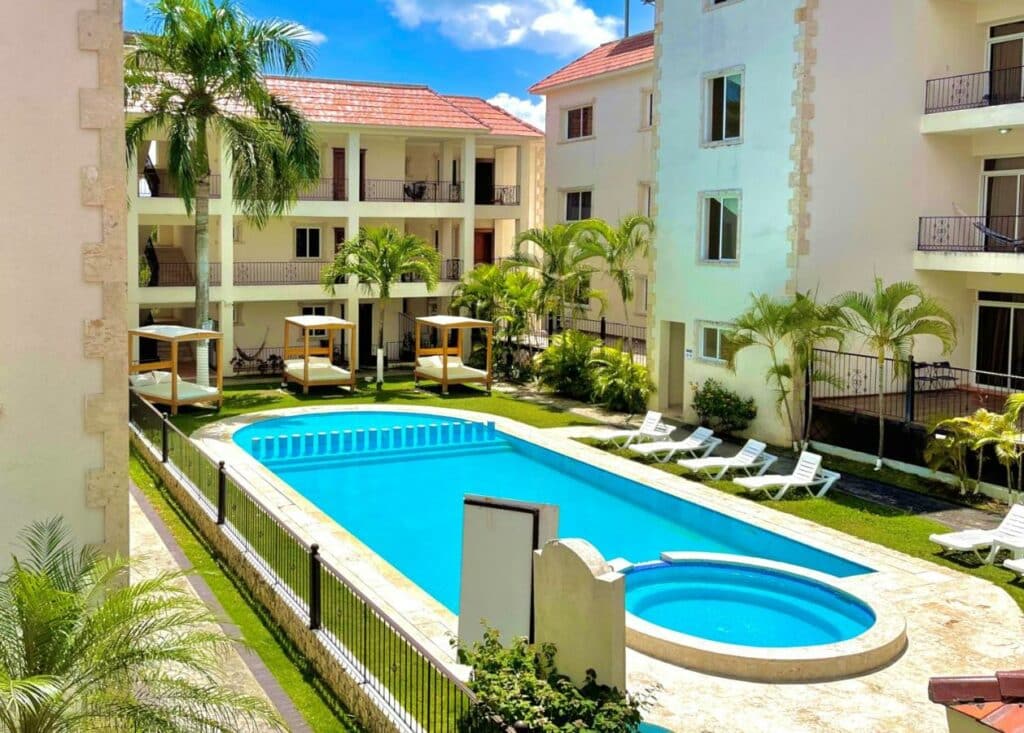Piscina do Punta Cana Seven Beaches vista de cima. Há diversas espreguiçadeiras e camas balinesas, que são estruturas relaxantes com colchão e cortinas nas laterais, espalhadas ao redor do local, que é rodeado pelos prédios do hotel.