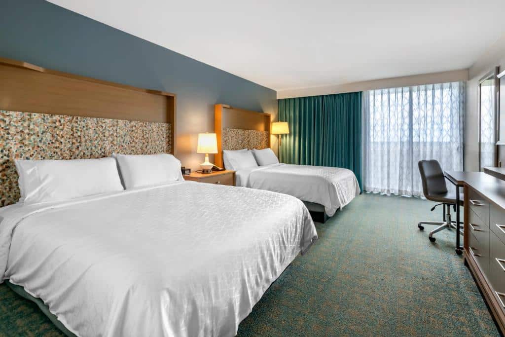 Quarto amplo do Holiday Inn Orlando – Disney Springs com duas camas de casal, uma varanda com cortinas e, em frente a cama, há uma cômoda com gavetas, ums mesa de escritório e uma cadeira giratória
