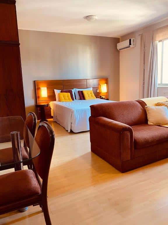 quarto do Apart-hotel Mercure Prinz com uma cama de casal no centro do quarto, além de um sofá em frente a cama e uma mesa de vidro no lado esquerdo da imagem, com duas cadeiras estofadas.