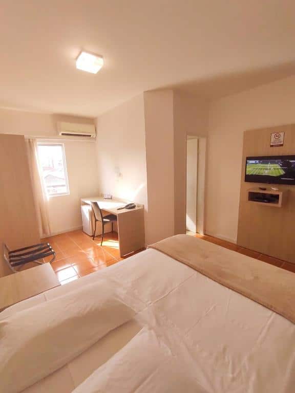 cama de casal em frente a uma televisão smart, com uma mesa para trabalho à esquerda do quarto no Avenida Palace Hotel em Joinville.