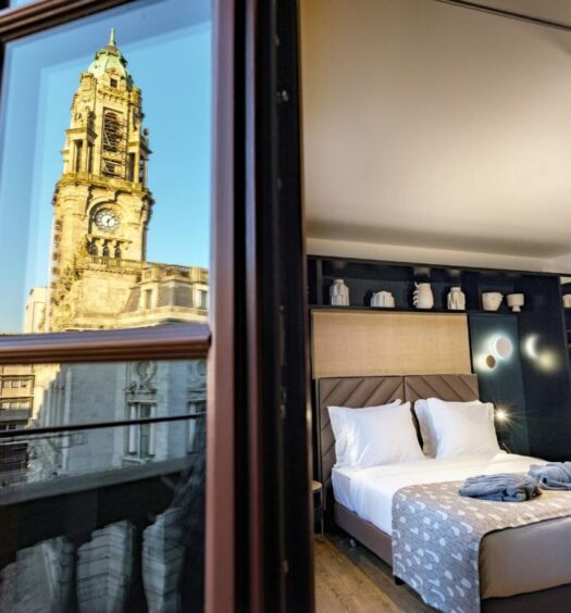 Vista do quarto do Axis Porto Club Aliados com cama de casal do lado direto, do lado esquerdo reflexo da Torre do Clerido no vidro da porta do quarto que dá acesso a sacada. Representa hotéis no centro do Porto.