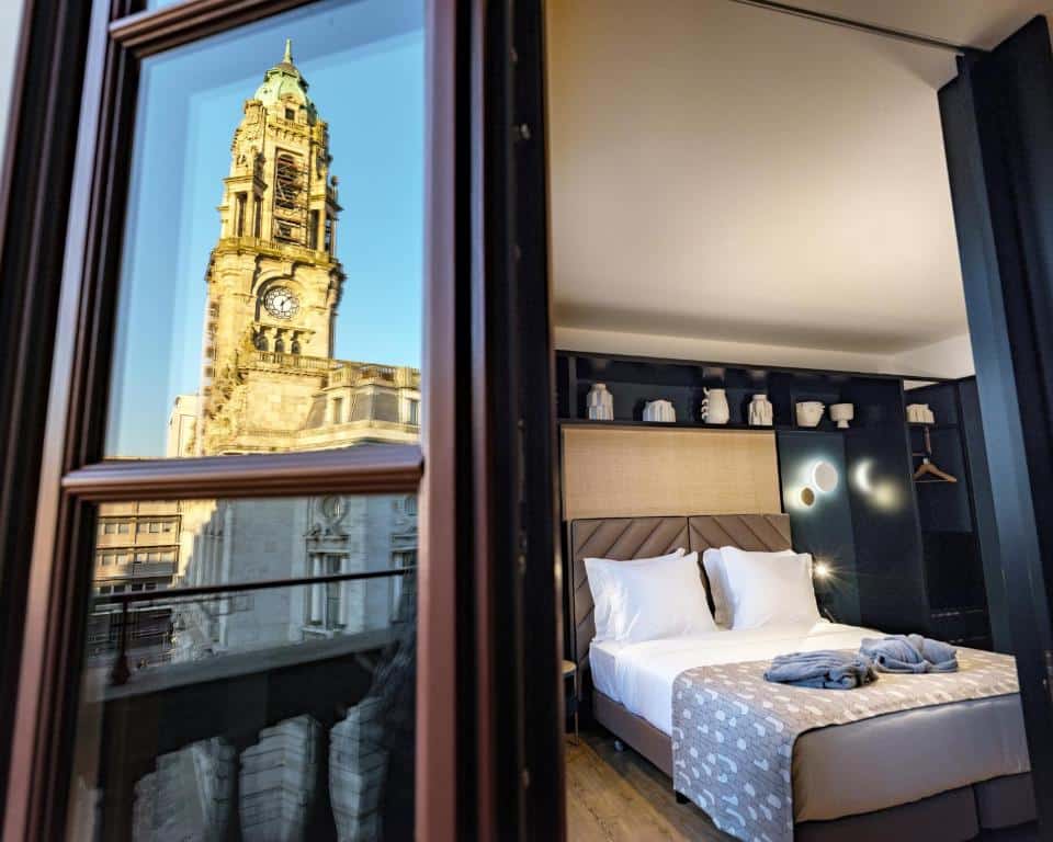 Vista do quarto do Axis Porto Club Aliados com cama de casal do lado direto, do lado esquerdo reflexo da Torre do Clerido no vidro da porta do quarto que dá acesso a sacada. Representa hotéis no centro do Porto.