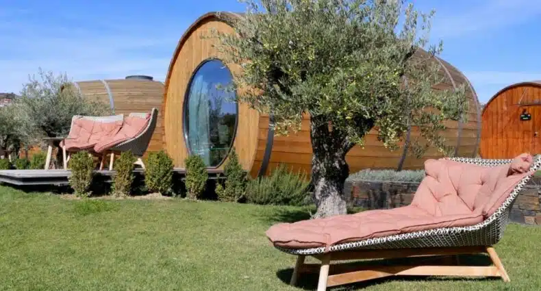 Vista da frente de um acomodação em forma de barril de vinho do The Wine House Hotel – Quinta da Pacheca, durante o dia com cadeira estofada a frente, atrás da cadeira uma arvore e atrás da arvore a hospedagem. Representa hotéis no Vale do Douro.