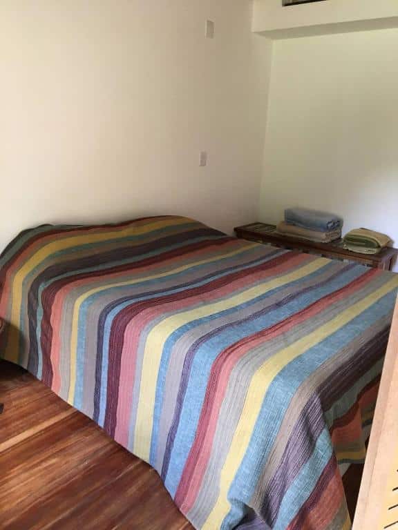 Uma foto de um quarto no Brandt Huss, para representar o post sobre pousadas em Santa Tersa (ES). Vemos uma cama de casal com lençóis listrados e coloridos sobre um piso de madeira.