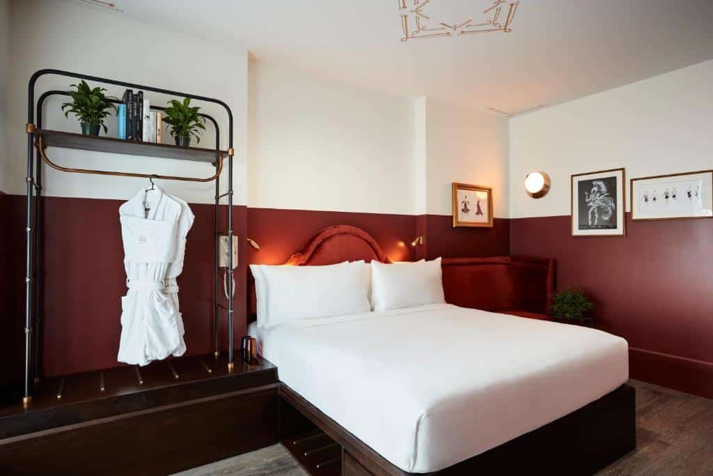 suíte retrô do CIVILIAN Hotel com uma cama de casal ao centro, um banco de canto de veludo vermelho ao lado direito da cama e do lado esquerdo um cabideiro de metal com dois roupões brancos pendurados.