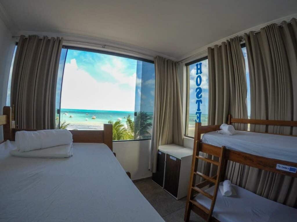 Quarto do Maraga Beach Hostel com duas beliches, um armário abaixo da janela, além de janelas com vista do mar azulado com barcos navegando. À direita, em uma janela de vidro, tem um adesivo escrito "hostel"