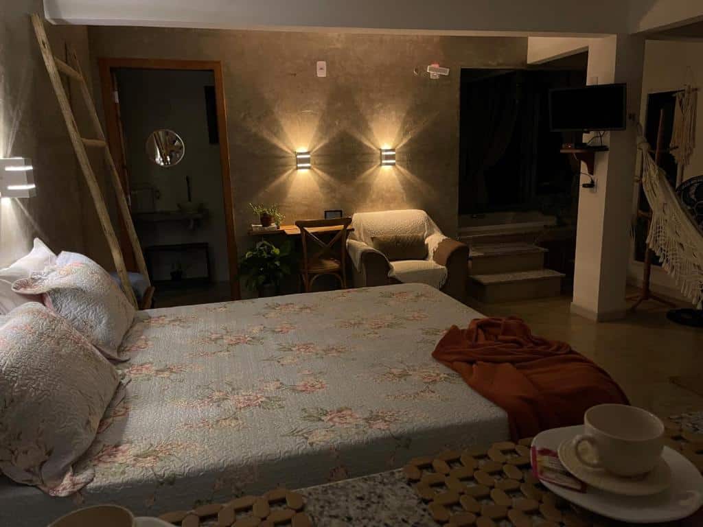 Quarto da Estância Shangri-La, representando o post sobre pousadas em Santa Teresa (ES). O ambiente é moderno e comporta uma cama de casal, no seu lado direito há uma porta para ao banheiro. Ao lado direito dessa porta há uma escrivaninha de madeira com uma cadeira e uma poltrona. Na frente da cama há um pilar e uma TV fixada nele. No canto inferior da foto, ao lado esquerdo da cama, vemos de relance uma bancada com uma xícara de porcelana em cima.