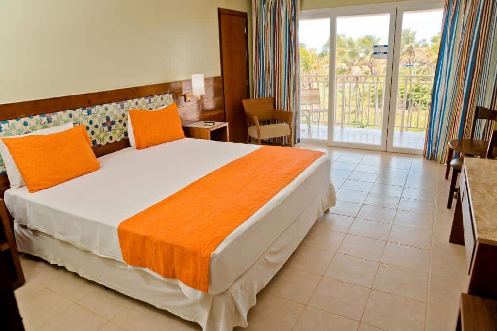 Quarto do hotel Gran Hotel Stella Maris Urban Resort, ilustrando o post de hotéis para o carnaval de Salvador. A cama está do lado esquerdo da suíte, e é box de casal. Do seu lado direito, há uma mesa de cabeceira e um abajur acima, também há uma porta no canto da parede e uma a poltrona. A janela da suíte está no fundo da foto, e é grande, iluminando bastante o ambiente. De frente para a casa, temos visão parcial de uma escrivaninha. O quarto é bem espaçoso.