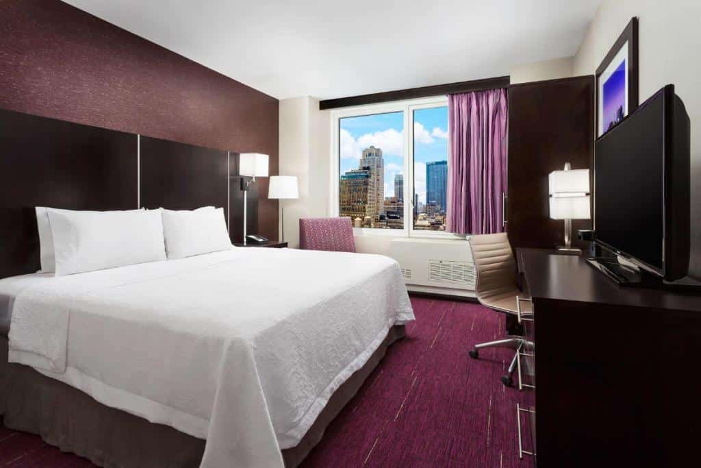 suíte do hotel Hampton Inn perto da Times Square Central com uma cama de casal no lado esquerdo do quarto e uma cômoda de madeira escura em frente, no lado direito, com uma televisão de tela plasma em cima.