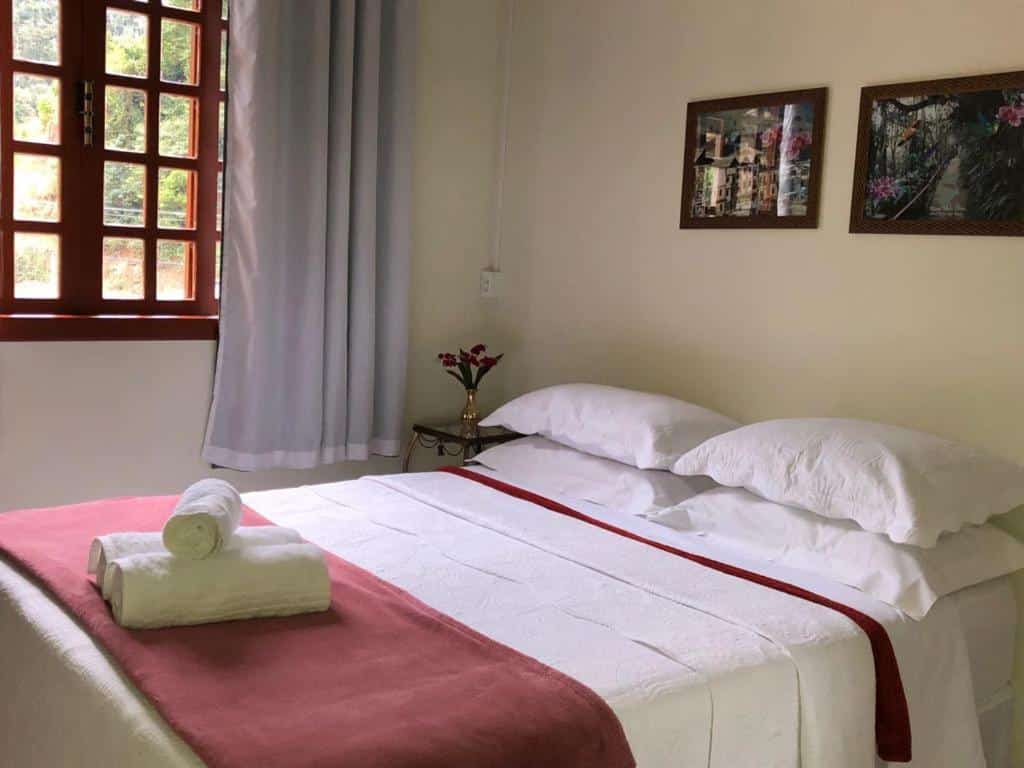 Quarto na Hospedaria Aconchego, para representar o post sobre pousadas em Santa Teresa (ES). Uma cama box de casal ocupa o maior foco da imagem, e ao seu lado esquerdo há uma pequena mesa de com um vaso de flor em cima. Ainda no lado esquerdo do quarto, também ao lado da cama, há uma janela.