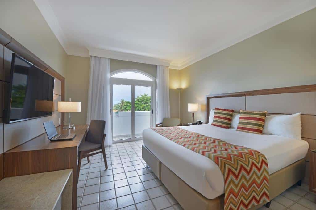 Quarto no Hotel Deville Prime Salvador, ilustrando o post de hotéis para o carnaval de Salvador. A cama é box de casal, e está no lado direito da foto. De ambos seus lados há uma mesa de cabeceira com abajur em cima. Na sua esquerda, há uma poltrona perto de uma porta de vidro, com uma varanda. Na frente da cama, ocupando o lado esquerdo da foto, há uma longa mesa de madeira com um notebook em cima e uma cadeira, e também há uma TV fixa em um painel.