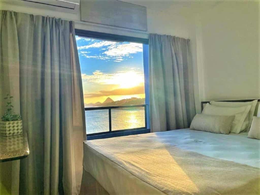 Quarto do Hotel Espadarte. Uma cama de casal no lado direito, de frente uma mesa. No lado esquerdo da cama, uma janela grande com cortina e vista do mar.