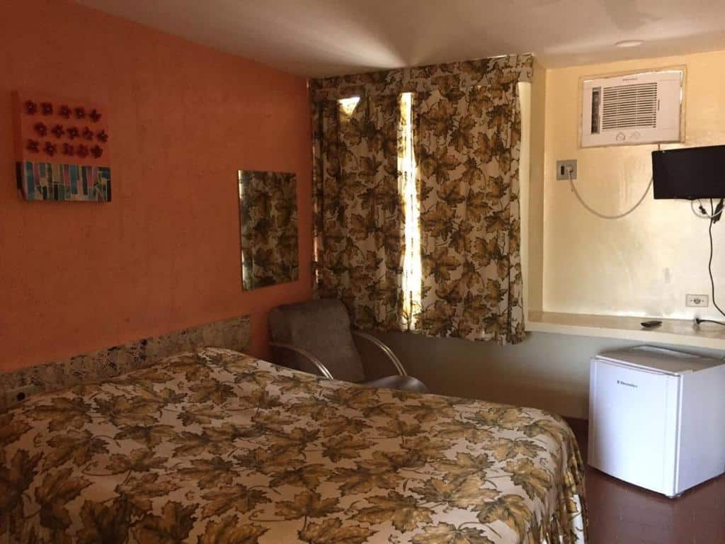 Um quarto no Hotel Pousada Itamaracá, para ilustrar o post sobre pousadas em Itamaracá (PE). Podemos ver a cama de casal, e do seu lado direito há uma poltrona e uma janela. No canto inferior direito da foto há um frigobar e uma TV suspensa na parede.