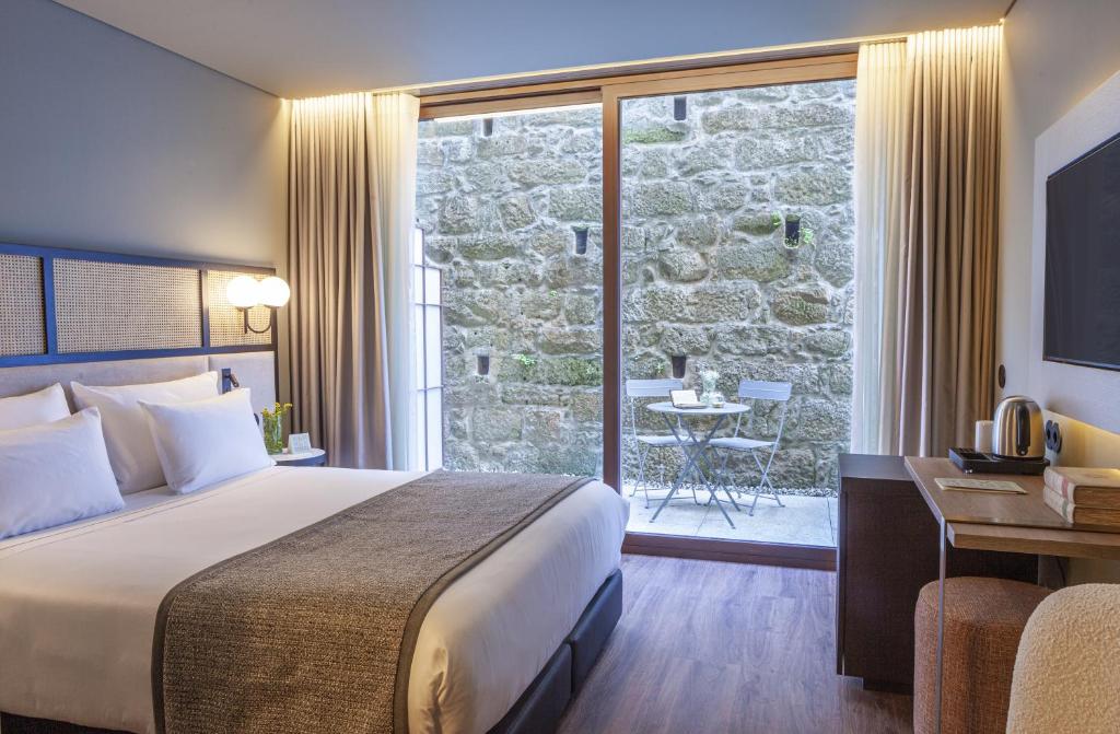 Quarto do Hotel das Virtudes com cama de casal do lado esquerdo da imagem, em frente a cama uma mesa de madeira e um frigobar ao lado da mesa, ao fundo uma porta de vidro que dá acesso a varanda. Representa hotéis no centro do Porto.