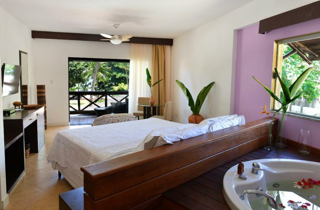 Foto do Iara Beach Hotel Boutique, ilustrando o post de hotéis para o carnaval de Salvador. A suíte é charmosa e extensa. A cama do quarto está de costas para a foto, e atrás dela, no canto inferior direito, vemos a hidro com pétalas de rosas que a suíte possui. Do lado da hidro, há uma janela. No lado esquerdo do quarto, ao lado da cama, há uma mesa com gavetas e algumas decorações em cima, e a TV está fixa na parede. No fundo do quarto, mais para a frente de onde está a cama, há uma porta larga com cortinas que dá acesso à varanda da suíte.