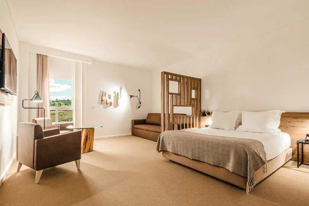 Quarto do Lamego Hotel & Life com cama de casal do lado direito da imagem, do lado esquerdo da cama uma área de estar com sofá uma mesa de centro redonda e duas poltronas. Representa hotéis no Vale do Douro.