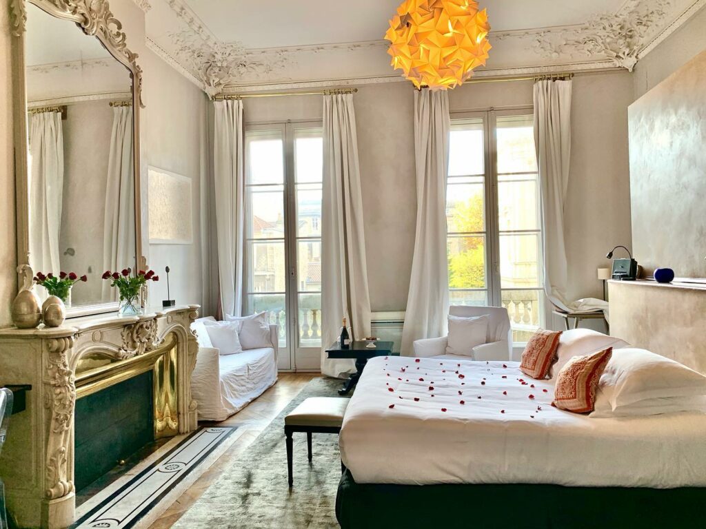 Quarto do BB L'Hôtel Particulier Bordeaux. Em primeiro plano existe uma cama de casal toda coberta por pétalas de rosas. Em frente está um balcão decorado e com um espelho grande. Ao fundo estão duas portas de vidro levando para a varanda. Em frente a ela (e ao lado da cama) está um sofá, uma mesinha e uma poltrona.
