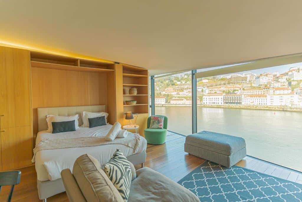 Quarto do LUXURY VIEWS by YoursPorto com cama de casal do lado esquerdo da imagem, do lado direito da cama uma poltrona e um banco estofado perto das janelas panorâmicas que tem vista do rio Douro.