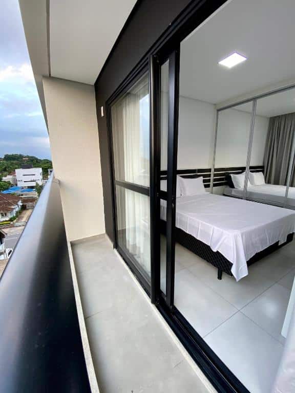 vista de dentro da sacada particular para o quarto da pousada MaxLoft em Joinville, onde é possível ver uma cama de casal com armários embutidos ao lado, e espelhos verticais cobrindo as portas de correr do móvel.