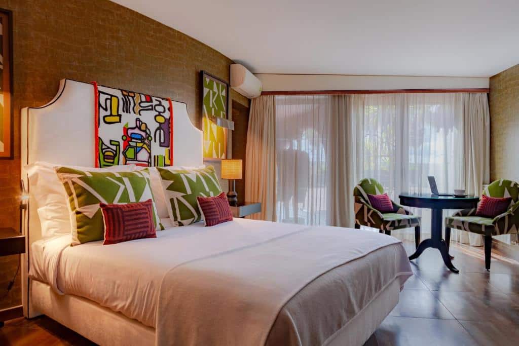 Quarto do hotel Montargil Monte Novo. A cama está posicionada no lado esquerdo do quarto e ao lado direito há uma mesa redonda com duas cadeiras e uma parede de vidro com longas cortinas. Imagem para ilustrar o post hotéis no Alentejo.