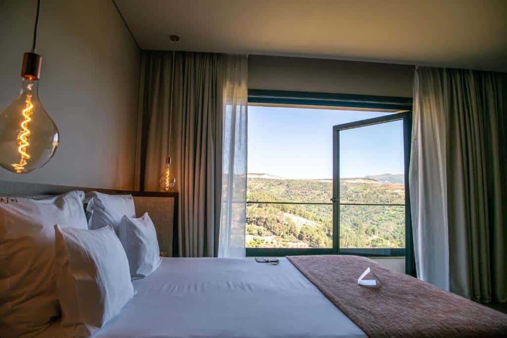 Quarto do MW Douro Wine & Spa Experience Hotel Collection com cama de casal do lado esquerdo da imagem a frente e do outro lado uma janela ampla com cortinas com vista para as montanhas. Representa hotéis no Vale do Douro.