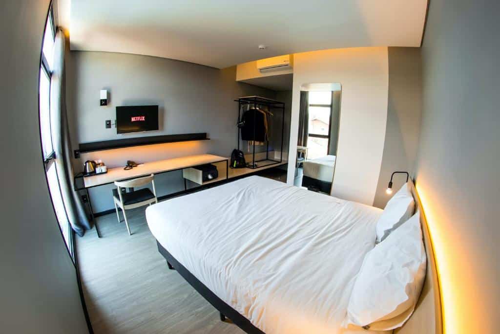 quarto do Naalt Hotel Joinville com uma cama de casal à direita, uma mesa de madeira em a cama, com uma televisão smart presa à parede, em cima da mesa. No lado direito da cama já um espelho vertical na parede.