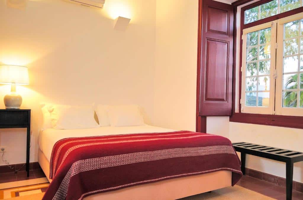 Quarto do hotel Parque de Natureza de Noudar. A cama está centralizada, no lado esquerdo da cama há uma mesinha com abajur e ao lado direito há uma janela. Imagem para ilustrar o post hotéis no Alentejo.