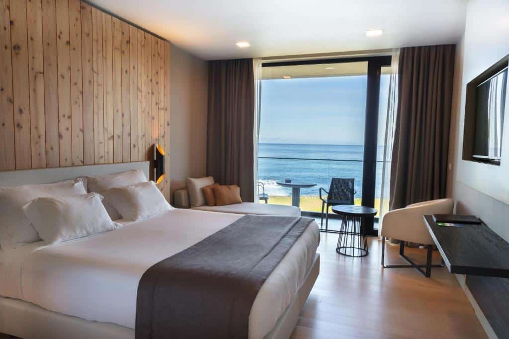 Quarto do Pedras do Mar Resort & Spa com cama de casal do lado esquerdo da imagem, em frente a cama uma TV pendurada na parede e do lado esquerdo da cama um sofá. Representa hotéis 5 estrelas em Portugal.