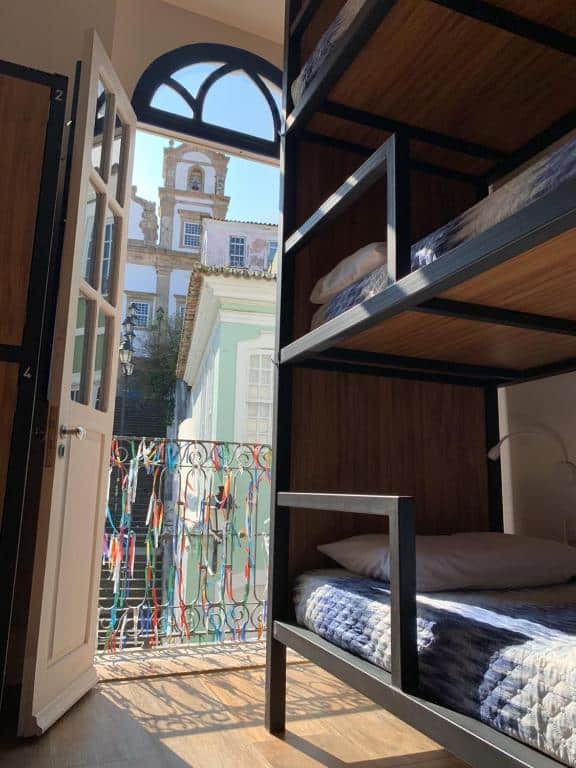 Foto de um quarto na Bahia Pelô Hostel. Na direita, vemos algumas beliches, e atrás delas há uma porta com uma varanda que dá vista para a rua de Pelourinho.