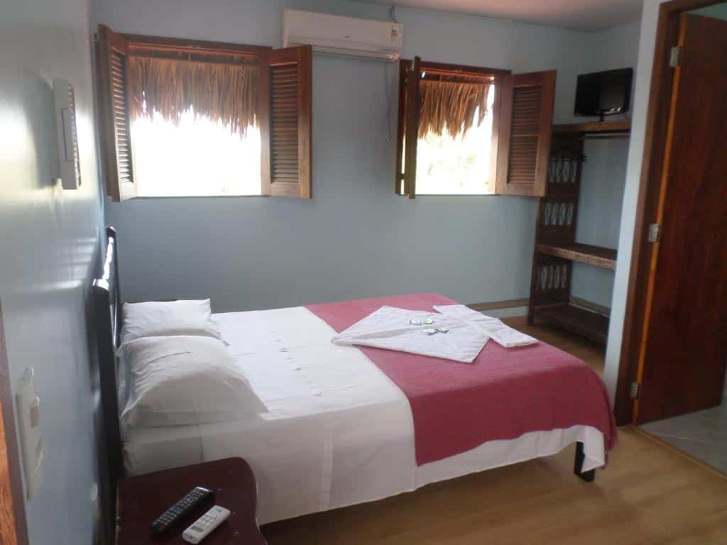 Quarto da Pousada Brasita com uma cama de casal com dois travesseiros, ao fundo duas janelas de madeira com um ar-condicionado na parede, no canto um espaço para colocar as bagagens e um tv em cima.