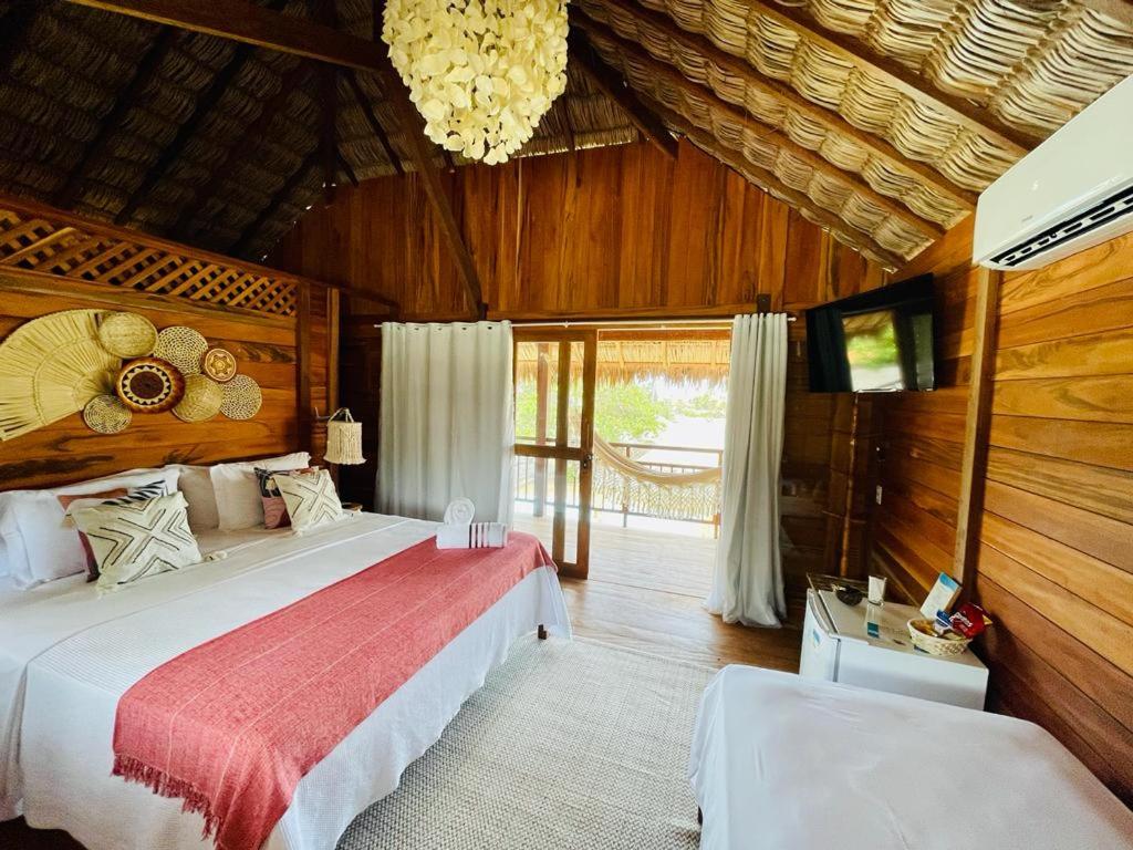Quarto da Pousada Casa Caju Bangalows com paredes de madeira, uma cama de casal grande com travesseiros e almofadas, em frente um frigobar, uma tv da parede e um ar-condicionado, e ao lado uma porta de vidro com cortinas brancas que dá acesso a varanda com uma rede.