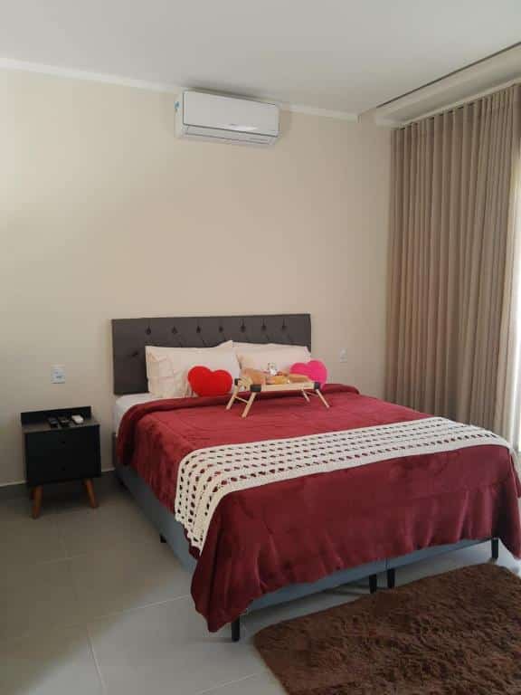 Quarto na Pousada Casa no Sítio, representando o post sobre pousadas em Santa Teresa (ES). Há uma cama box de casal, e em cima dela tem uma bandeja com amenidades da pousada. Do lado direito da cama há uma janela com cortina bege, e do lado esquerdo há uma mesa de cabeceira com controles remotos em cima.