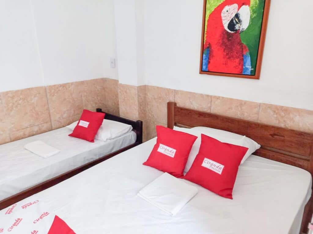 Um quarto na Pousada Ciranda, para representar o post sobre pousadas em Itamaracá (PE). Vemos uma cama de casal, com almofadas com o logo da pousada em cima, e outra cama de solteiro ao seu lado esquerdo. Na parede, acima da cama de casal, há um quadro com uma arte de uma arara vermelha.