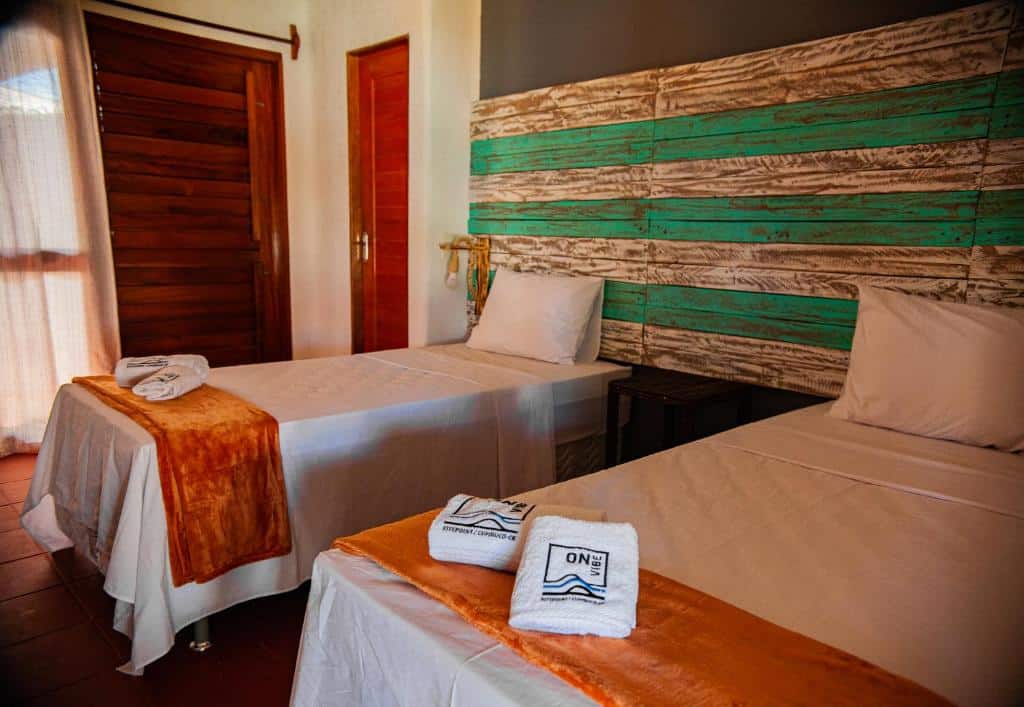 Quarto da Pousada ON VIBE Cumbuco com dus camas de solteiro, uma mesinha separando as duas e ao lado esquerdo da foto há duas portas de madeira e uma cortina branca.