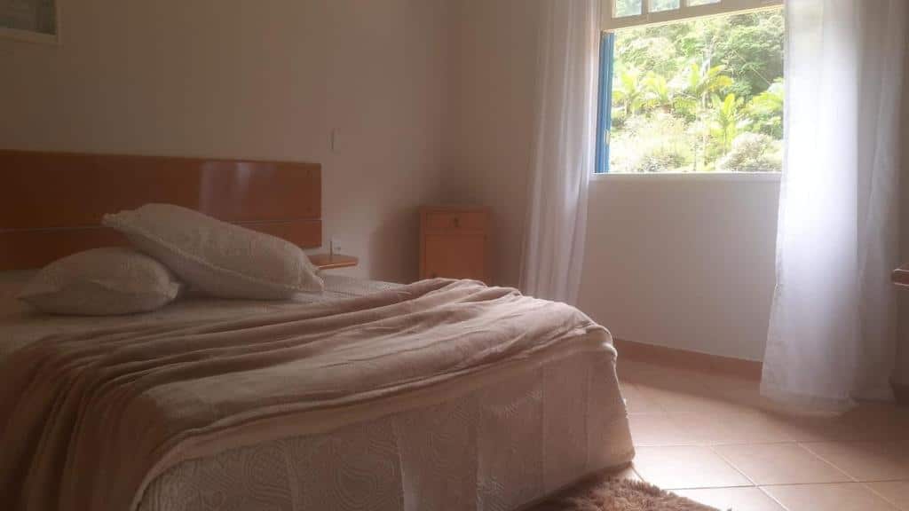 Quarto na Pousada Paradiso, para ilustrar o post sobre pousadas em Santa Teresa (ES). No canto esquerdo temos a visão de uma cama de casal, e ao seu lado direito há uma pequena mesa de cabeceira de madeira, encostada na parede. Ainda no lado direito do quarto, há uma janela que areja o ambiente.