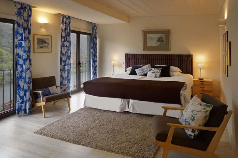 Quarto do Quinta de la Rosa com cama de casal no centro do quarto, com uma cômoda em cada lado da cama com luminária em cada lado da cama a frente uma poltrona estofada. Representa hotéis no Vale do Douro.
