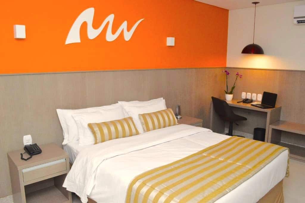 Quarto no Real Classic Bahia Hotel, ilustrando o post de hotéis para o carnaval de Salvador. A parede na qual a cama está encostada é laranja e possui um logo. A cama é box de casal e possui uma mesa de cabeceira em ambos os lados. Uma escrivaninha com um notebook, cadeira, telefone, vaso decorativo e um lustre em cima está do lado direito da cama, encostada na parede.