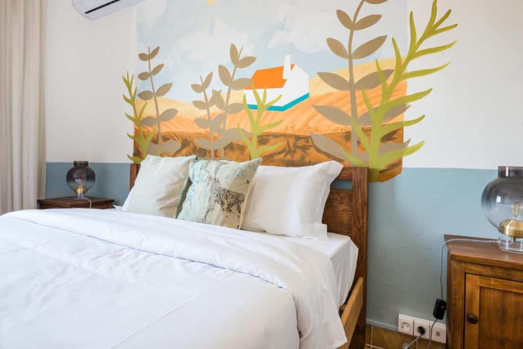 Quarto do hostel Selina Milfontes. A cama de casal está centralizada, contém dois travesseiros e duas almofadas em cima e também há mesinhas com abajures em cada lado da cama.