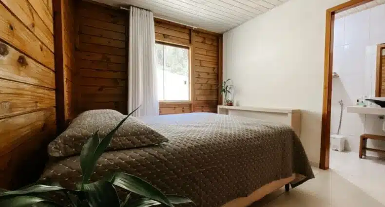 Um quarto em Sítio Ancestrale, em Santa Teresa (ES). Há uma cama box de casal encostada na parede, essa que é feita de madeira, enquanto as outras paredes da suíte são brancas. Do lado esquerdo da cama, vemos parcialmente um vaso de planta. Já do lado direito, está a janela do quarto com uma cortina branca. A parede de frente para a cama possui uma pequena mesa branca com um vaso de planta em cima, e do lado dela há uma porta para o banheiro do quarto.