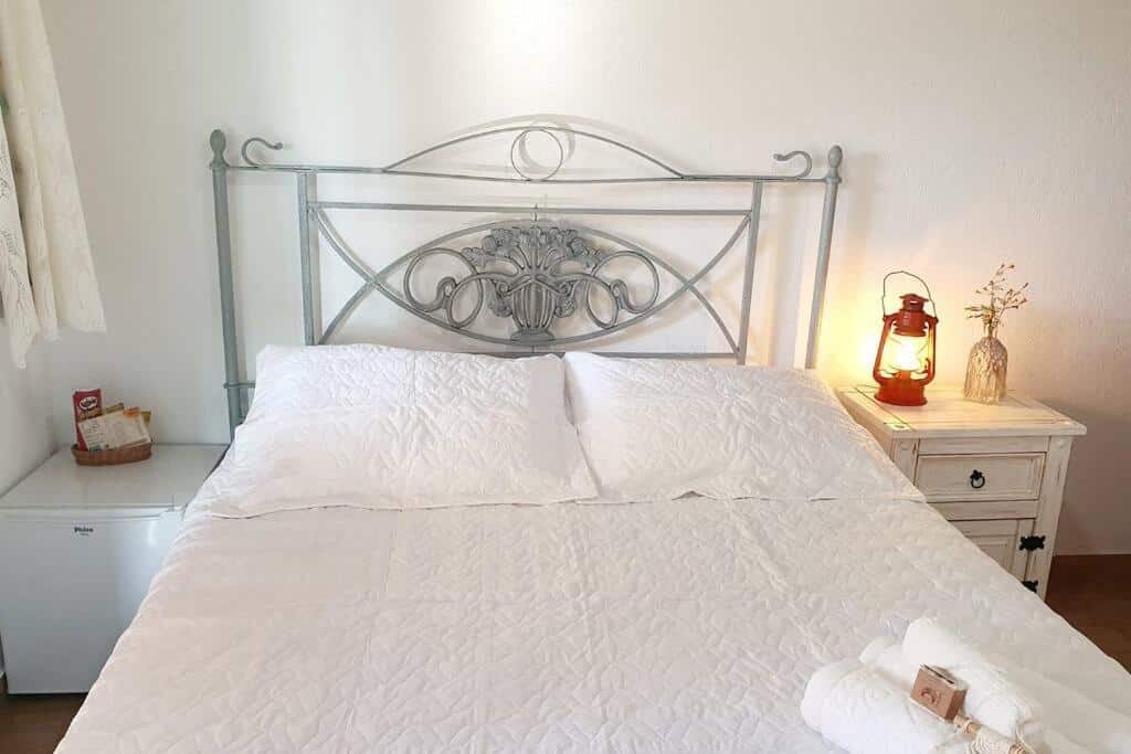 Quarto no Sítio Juara, representando o post sobre pousadas em Santa Teresa (ES). Uma cama de casal está no centro da foto, com lençóis brancos. Do seu lado direito há uma mesa de cabeceira branca com um abajur em cima e vaso com flores, e no lado esquerdo há um frigobar com amenidades, além de ter também a janela do quarto.