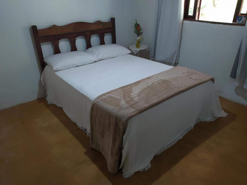 Quarto no Sítio Recanto Amado, para ilustrar o post sobre pousadas em Santa Teresa (ES). Há uma cama de casal, e ao seu lado direito há uma pequena mesa com um vaso de flor em cima e uma janela na parede.