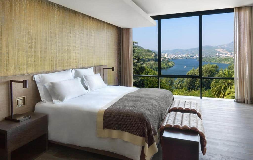 Quarto do Six Senses Douro Valley, com cama de casal do lado esquerdo da imagem com um baú no pé da cama e do lado esquerdo da cama janelas panorâmicas com vista para o rio. Representa hotéis no Vale do Douro.