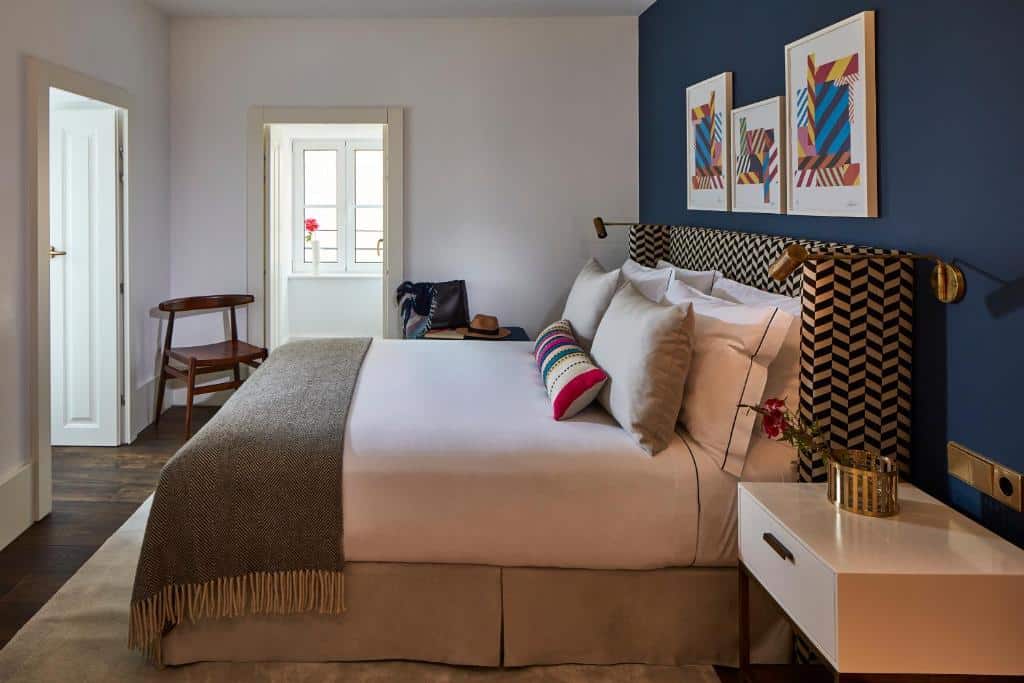Quarto do The Lumiares Hotel & Spa com uma ampla cama de casal com travesseiros e almofadas, do lado esquerdo do quarto há duas cadeiras de madeira, na parede atrás da cama há três quadros pendurados, assim como luminárias presas em direção da cama, para representar hotéis bem localizados em Lisboa