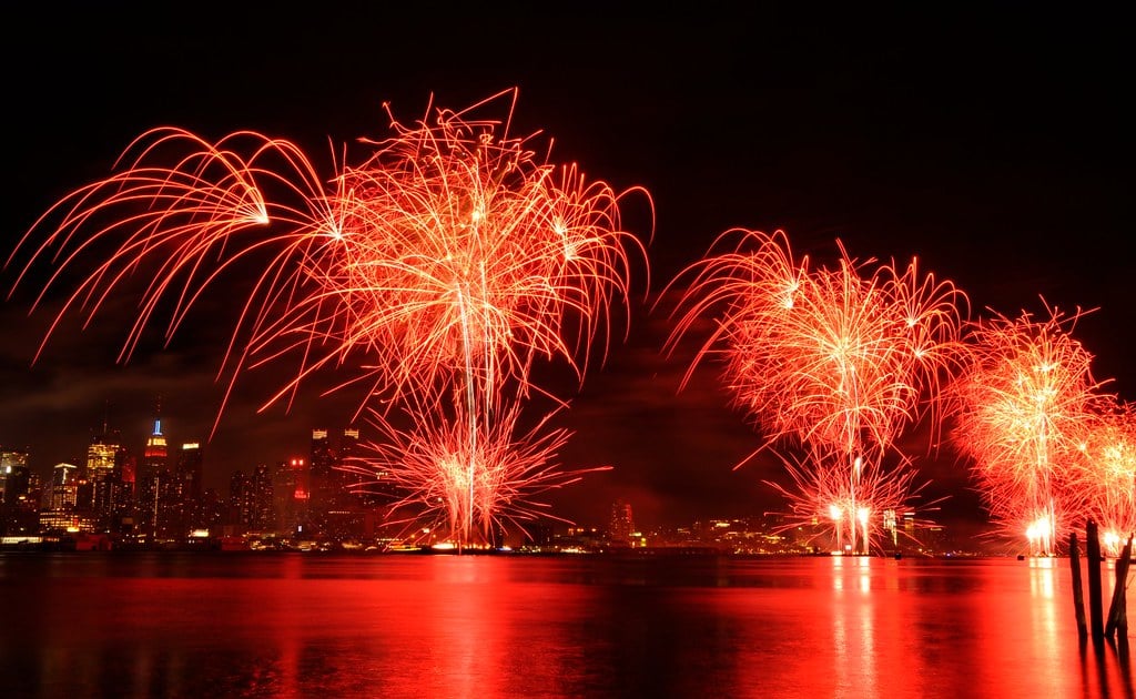 Fogos de artíficio vermelhos sendo lançados na cidade de Nova York e relfetindo suas luzes no rio