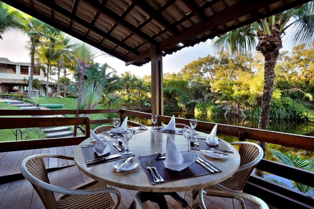 Área de restaurante do hotel Deville Prime Salvador. Vemos uma mesa redonda posta com talheres, taças e lenços, e com quatro cadeiras ao seu redor. Está no canto de uma varanda, e vemos uma bela vegetação e um lago. É um ambiente que passa uma sensação relaxante e de frescor.