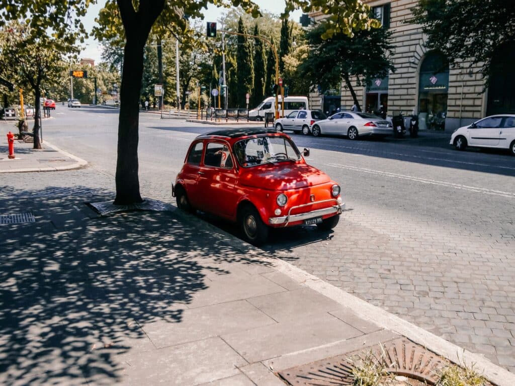 vista de carros nas ruas amplas de Roma, há várias árvores ao redor e prédios com fachada de tijolo